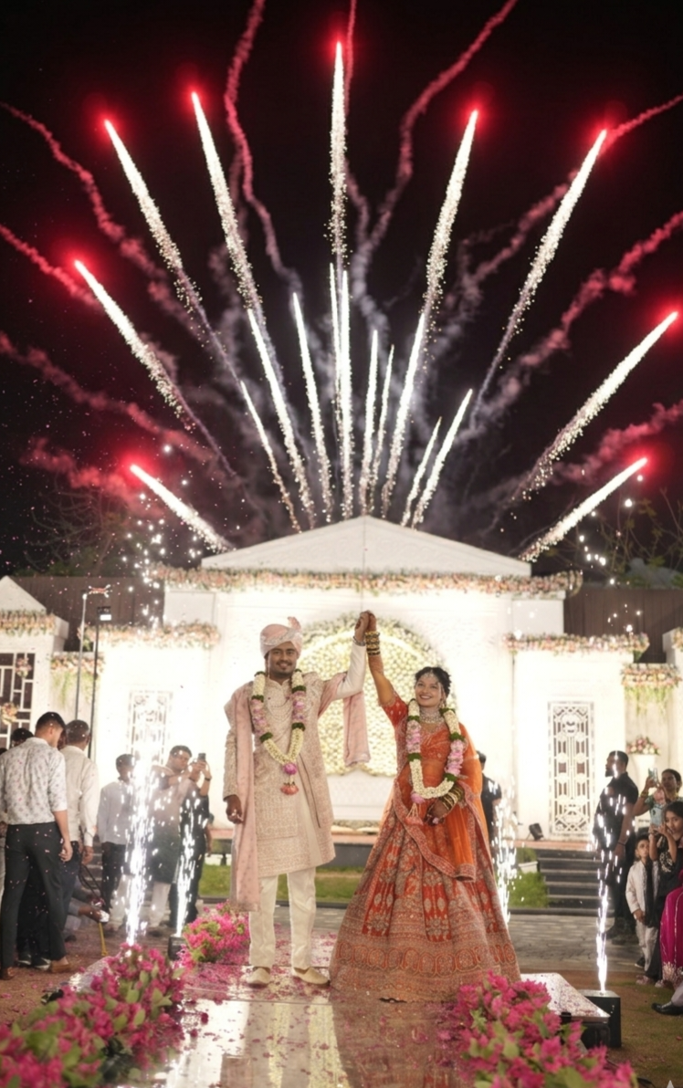 Bride and groom raising joined hands as fireworks burst above a white floral wedding stage